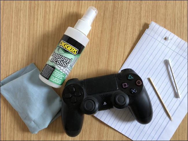 Контроллер DualShock 4 находится на листе бумаги рядом с зубочисткой, ватной палочкой, распылителем изопропилового спирта и тканью. 
