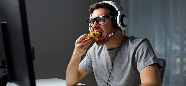 Mężczyzna siedzący przed komputerem, ubrany w słuchawki i je pizzę.