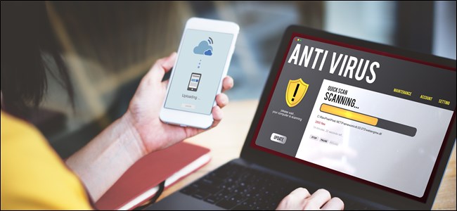 Una mano que sostiene un teléfono inteligente que ejecuta un antivirus, junto a una computadora portátil en un escritorio que también ejecuta un análisis antivirus.